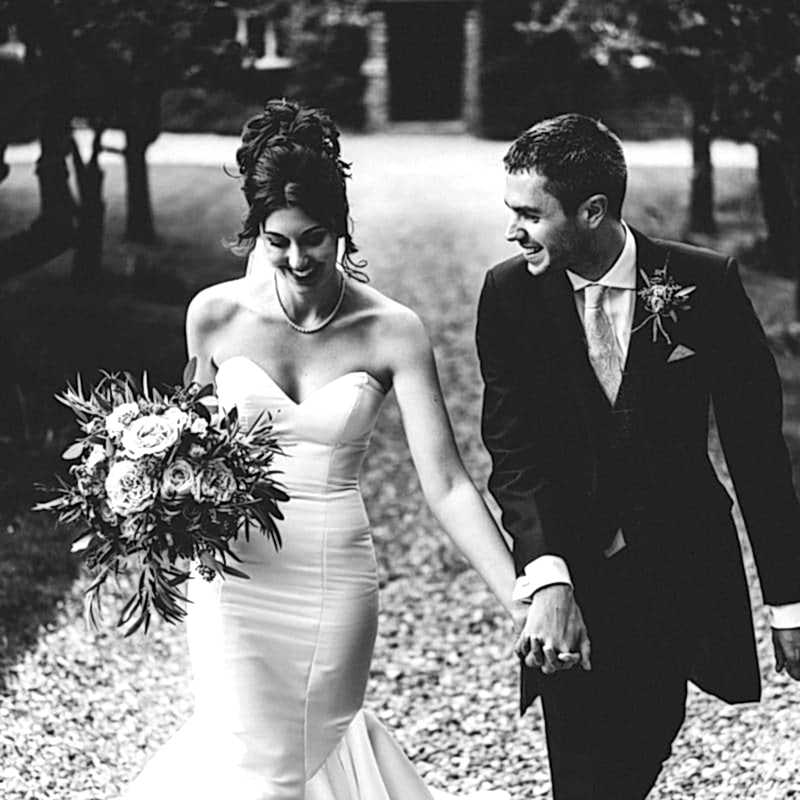 A bride and groom walk hand in hand on a gravel path, wearing formal wedding attire. The bride carries a bouquet and both are smiling, with trees and blurred background—capturing the elegant essence of Dodmoor House Wedding Photography.