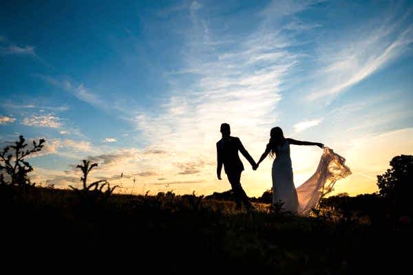 Dodford Manor Wedding Photographer captures the heartfelt moment of a bride and groom holding hands in a picturesque field at sunset.