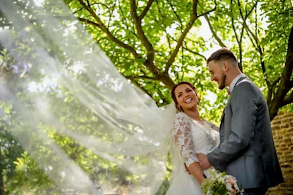 A bride and groom standing under a tree with their veil blowing in the wind, captured by Dodford Manor Wedding Photographer.