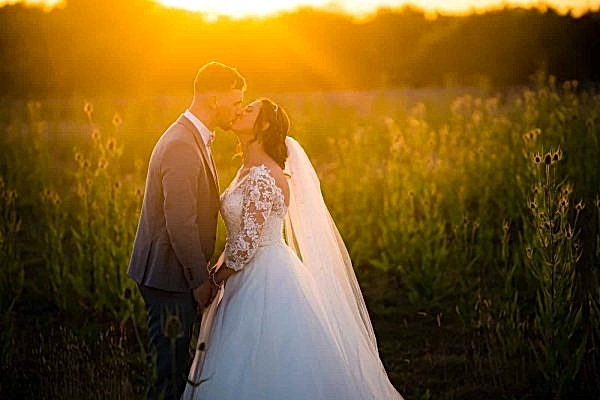 Dodford Manor Wedding Photographer captures a stunning moment of a bride and groom sharing their first kiss as husband and wife in a breathtaking field at sunset.