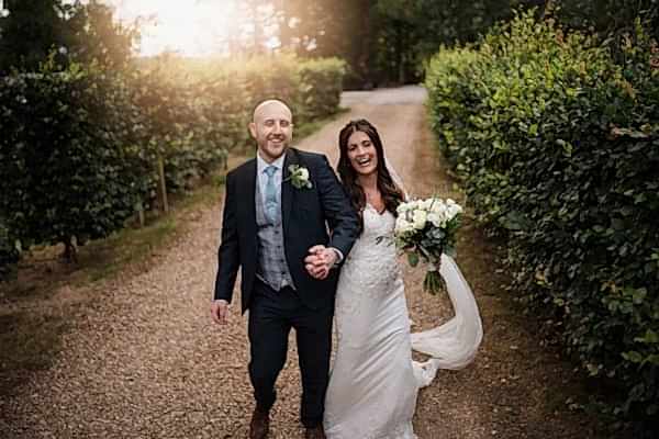 A Dodford Manor Wedding Photographer captures the enchanting moment of a bride and groom walking down a path in a garden.