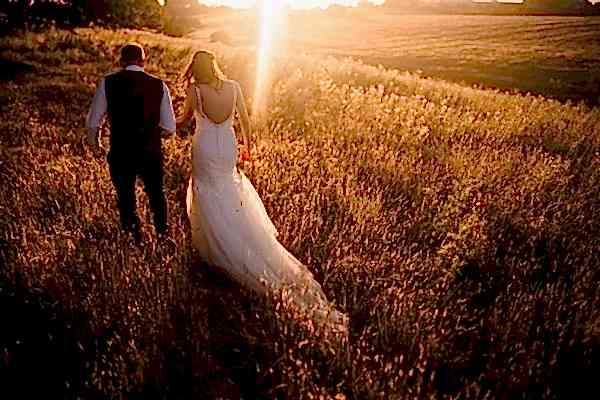 A Dodford Manor wedding photographer captures a beautiful moment as the bride and groom walk through a field at sunset.