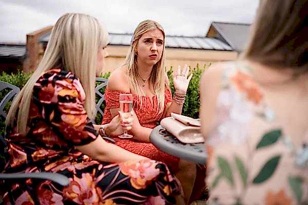 Dodford Manor Wedding Photographer capturing three women sitting at an outdoor table engaged in lively conversation.