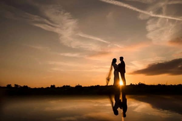 A bride and groom, captured by the Dodford Manor Wedding Photographer, standing in front of a serene pond at sunset.