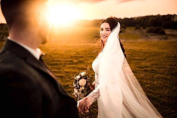 A Dodford Manor wedding photographer captures the intimate moment of a bride and groom holding hands in a breathtaking field at sunset.