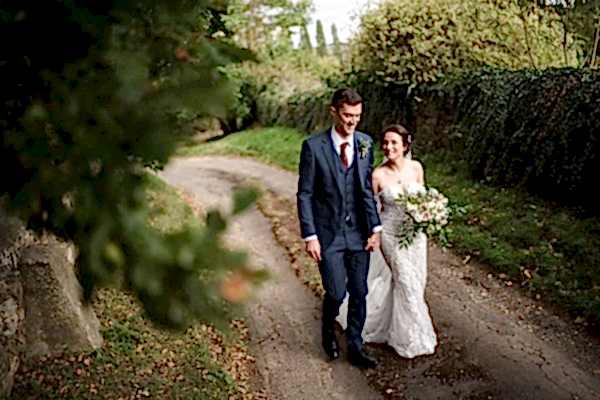 A Dodford Manor Wedding Photographer captures a beautiful moment of a bride and groom walking down a dirt path.
