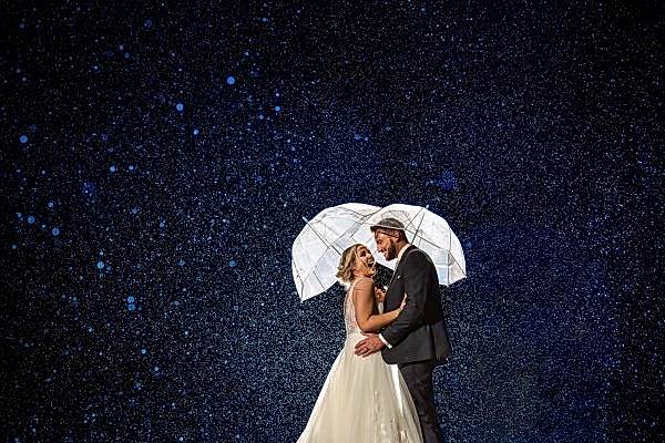 A bride and groom standing under an umbrella in the rain, captured by a Dodford Manor Wedding Photographer.