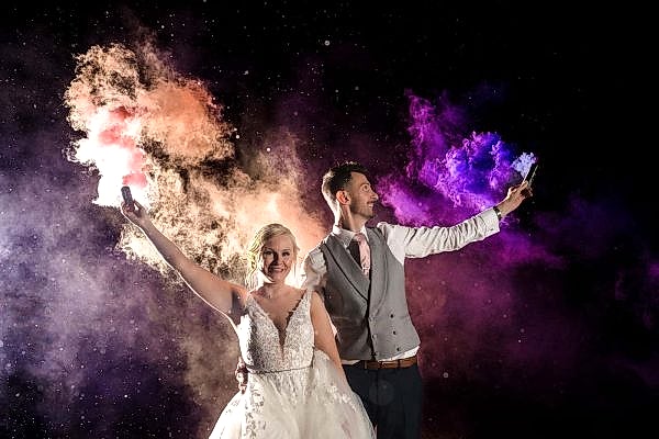 A Dodford Manor wedding photographer captures a bride and groom posing with colored powder in front of a dark background.