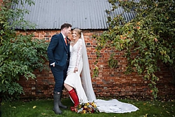 A Dodford Manor Wedding Photographer captures a bride and groom posing in front of a brick wall.