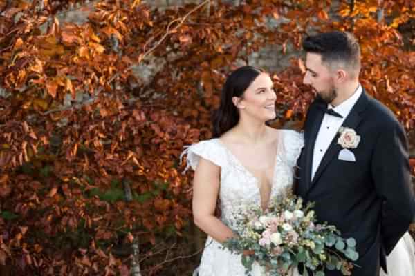 A Dodford Manor Wedding Photographer captures a bride and groom standing in front of a tree in autumn.