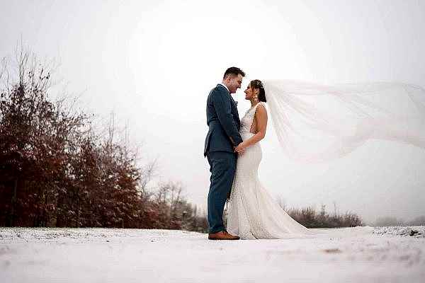 A bride and groom, captured by a Dodford Manor Wedding Photographer, standing in the snow with their veil blowing in the wind.