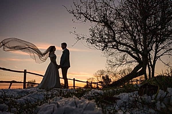A bride and groom standing in the snow with their veil blowing in the wind captured by a Dodford Manor Wedding Photographer.