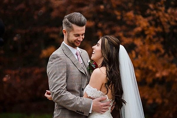 A bride and groom embracing in front of a tree, captured beautifully by a Dodford Manor Wedding Photographer.