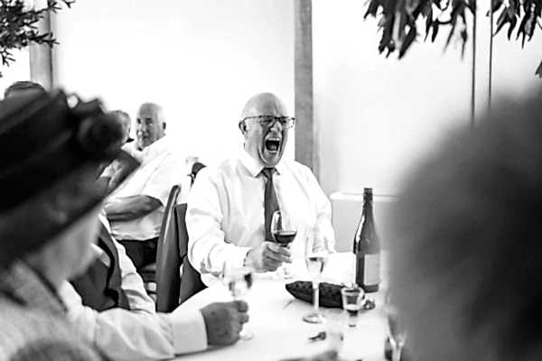 A Dodford Manor Wedding Photographer captures a black and white photo of a man laughing at a table.