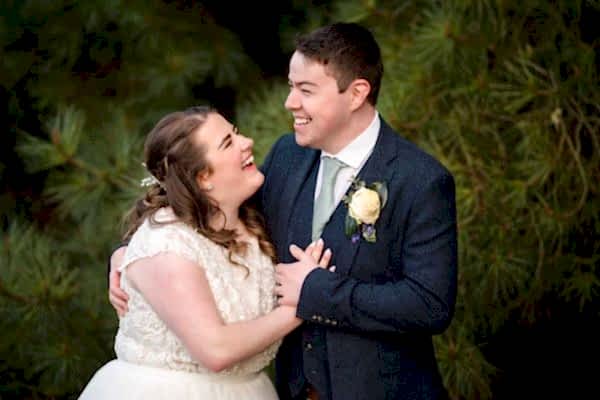 A bride and groom, captured by a talented Dodford Manor Wedding Photographer, share a joyous moment as they laugh together in front of a majestic pine tree.