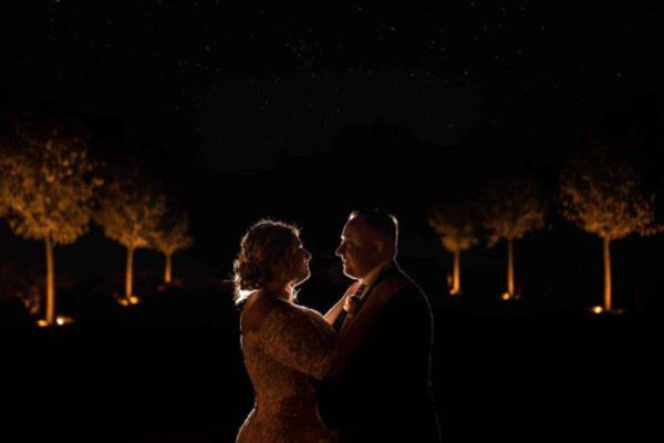 A bride and groom posing elegantly in front of trees at night, captured by a skilled Dodford Manor wedding photographer.