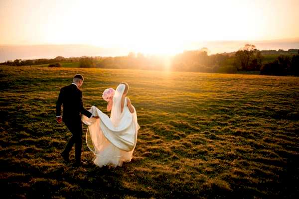 A bride and groom walking in a field at sunset captured by a Dodford Manor Wedding Photographer.
