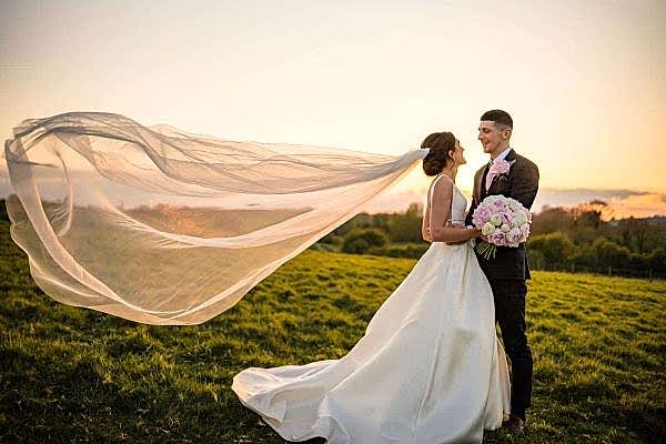 Dodford Manor Wedding Photographer captures a mesmerizing moment of a bride and groom standing in a field with their veil blowing in the wind.