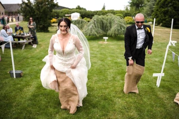 A bride and groom, captured by the skilled eye of a Dodford Manor wedding photographer, stroll gracefully through a lush garden landscape with sacks in hand.
