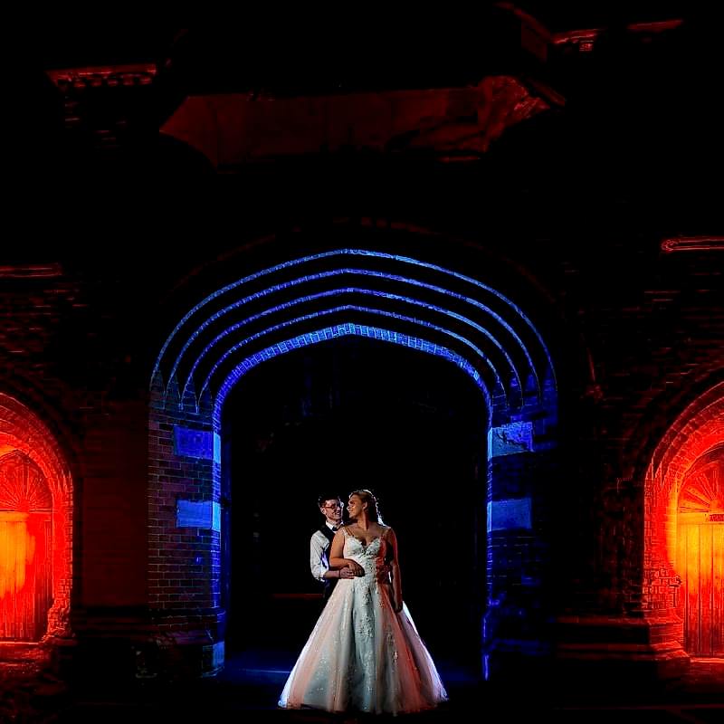 A bride and groom stand embraced under a gothic archway with dramatic red and blue lighting illuminating the scene, capturing a timeless moment of Holmewood Hall Wedding Photography.
