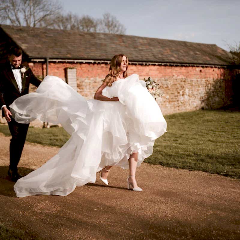 A bride in a white gown walks on a path in front of a rustic building, holding her dress, while a groom in a tuxedo follows closely, assisting with the gown's train.