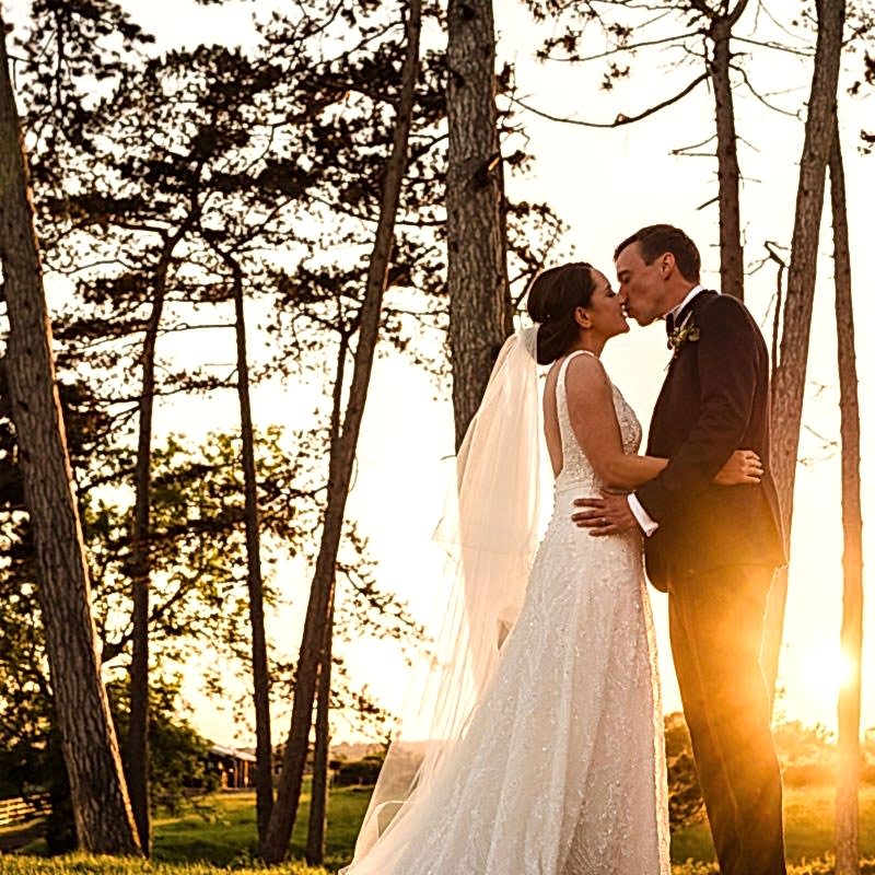 A couple in wedding attire shares a kiss in front of tall trees with sunlight streaming through, creating an ethereal glow in a natural outdoor setting, beautifully captured by a Leicestershire wedding photographer.