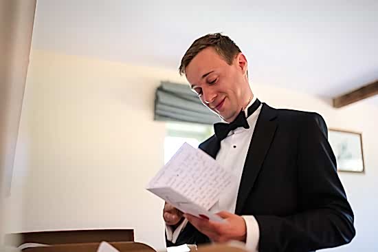 A person in a tuxedo reads a letter, likely penned by one of two wedding photographers from Buckinghamshire, standing in a room with beige walls and a green curtain.