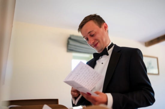 In a brightly lit room, a man in a tuxedo smiles as he reads a letter, capturing one of those unforgettable moments that showcase the benefits of two wedding photographers.