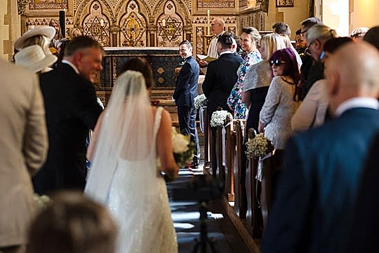A bride gracefully walks down the aisle in a church adorned with ornate decorations, two wedding photographers from Warwickshire capturing every moment, while the groom beams at the altar amidst seated guests.