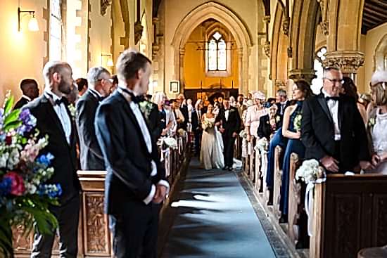 The bride and groom gracefully walk down the aisle of a Cambridgeshire church, surrounded by standing guests in formal attire, beautifully captured by the benefits of two wedding photographers ensuring every cherished moment is immortalized.
