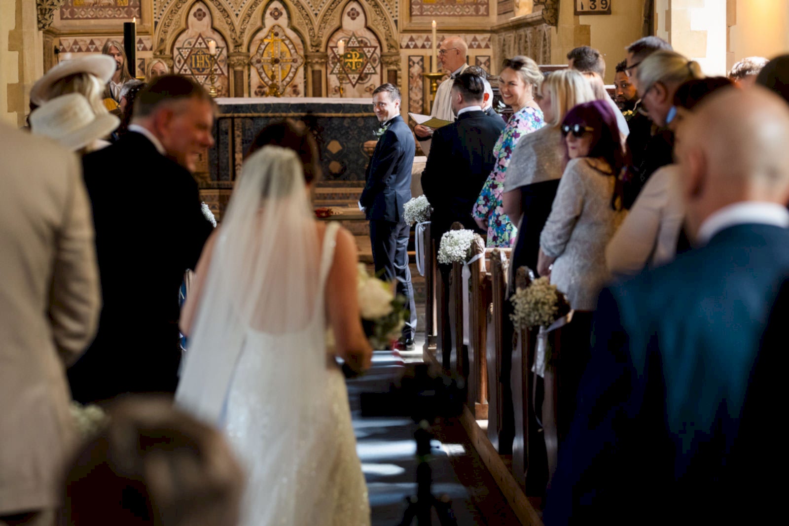 As the bride gracefully walks down the aisle in a charming church, guests look on with warm smiles, while two wedding photographers from Bedfordshire elegantly capture each breathtaking moment as the groom waits eagerly at the altar.