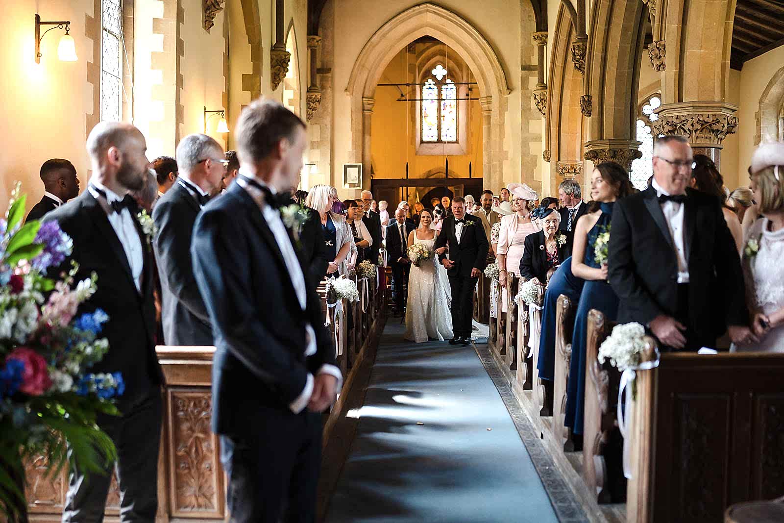 Bride walking down the aisle in a church, guests seated and standing, with two wedding photographers based in Leicestershire capturing every moment alongside two men in suits in the foreground.