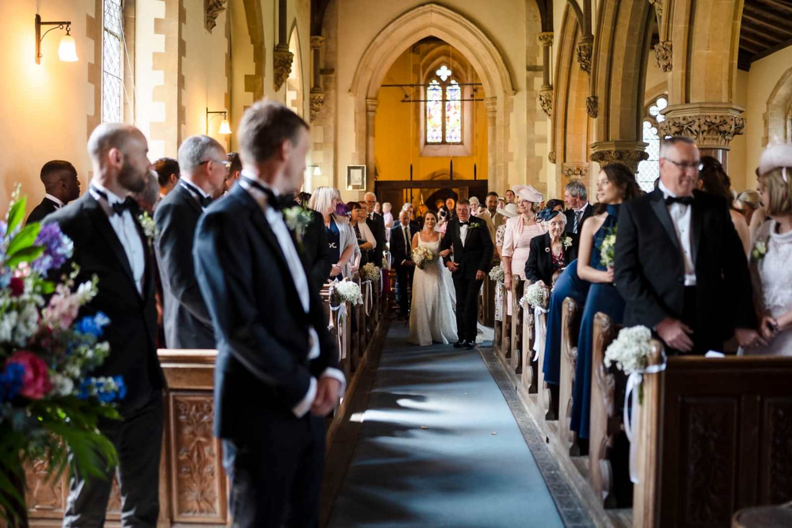 A bride walks down the aisle with her father in a church filled with guests, as two wedding photographers from Bedfordshire discreetly capture each magical moment.